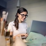 A smiling young woman wearing black glasses working comfortably on a laptop at a bright desk.