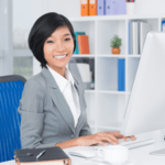 A smiling female administrative assistant in a grey suit working at a computer desk with organized office shelving in the background.