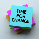 A stack of colorful square sticky notes viewed from above, with the top blue note displaying the handwritten text "TIME FOR CHANGE".
