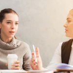 Two female colleagues seated together engaging in a focused discussion over coffee and notebooks.