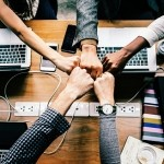 An overhead view of a group of colleagues bringing their fists together in a central fist bump over a wooden desk with laptops.