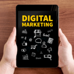 Two hands holding a black tablet against a wooden surface, displaying the text "DIGITAL MARKETING" in yellow alongside white icon doodles.