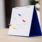 A white desktop flip calendar with a blue stand sitting on a wooden desk, marked with multi-colored pushpins sticking out from different dates.