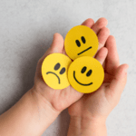 Two open hands holding three yellow circular cutouts displaying neutral, sad, and happy facial expressions against a neutral background.