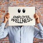 A person in a light blue dress shirt holding a sign over their face against a brick wall background; the sign displays cartoon googly eyes and the handwritten text "EMPLOYEE WELLNESS".