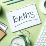 A top-down flat lay on a light green surface featuring a spiral notebook with the word "EVENTS" handwritten in black marker, surrounded by an analog alarm clock, keyboard, coffee cup, and stationery.