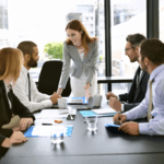 A female professional standing at the head of a conference table, leading and facilitating a meeting with a group of seated colleagues.