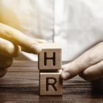 Hands placing and stabilizing two wooden blocks stamped with the letters "H" and "R" stacked on top of each other on a dark wooden surface.