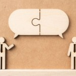 Wooden cutouts of two figures in ties under a large speech bubble puzzle piece, symbolizing effective communication and dialogue.