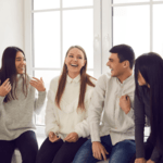 A diverse group of four young adults sitting together in a bright room, laughing and engaging in casual conversation near a large window.