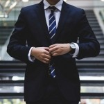 A sharp-looking businessman in a dark navy suit adjusting his necktie and jacket button inside a modern office lobby.