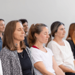 A group of people sitting together in a bright room with eyes closed during a guided mindfulness or meditation session.