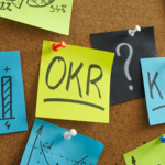 Brightly colored sticky notes pinned to a corkboard, with a central yellow note reading "OKR" surrounded by notes with charts and a question mark.