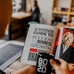 A person holding an open magazine featuring Richard Branson and the title "THE ENTREPRENEUR'S GUIDE TO SUCCESS & BUSINESS GROWTH" while enjoying a cup of coffee in a cafe.