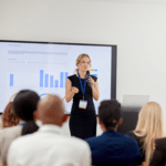 A blonde female speaker wearing a black dress and lanyard holds a microphone while delivering a presentation with data charts in front of an audience.