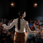 View from behind a female speaker with outstretched arms addressing a large audience from a dimly lit stage under overhead spotlights.
