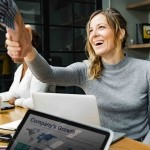 A smiling female professional leaning across an office table to shake hands with a colleague during a business meeting.
