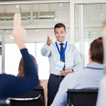A smiling corporate trainer in a shirt, tie, and lanyard gestures warmly toward an audience member raising their hand in a workshop.